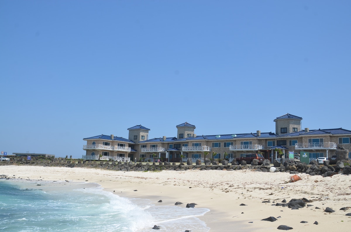 A beachfront view features a series of coastal buildings with blue roofs situated along a sandy shore. The gentle waves lap against the beach, highlighting the serene coastal environment. Rocky formations border the sand, enhancing the connection between land and sea.
