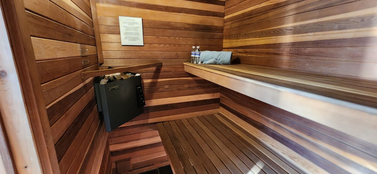 A sauna is featured with wooden paneling on the walls and benches. A small safe is positioned on the side, with two water bottles resting on a shelf. Soft lighting enhances the natural tones of the wood.