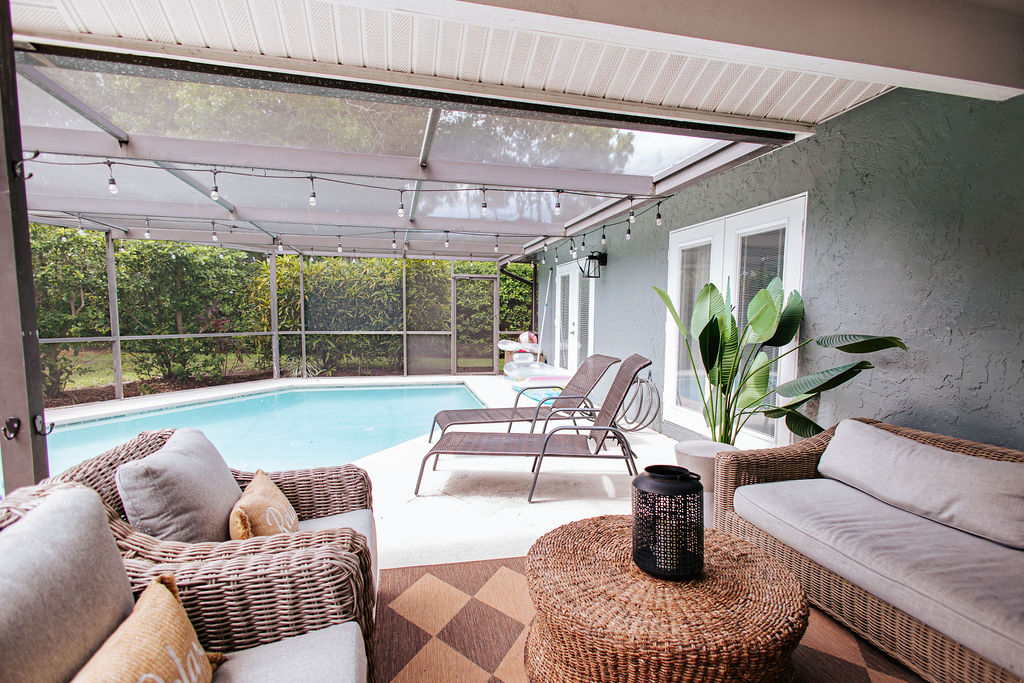 A serene outdoor area features a screened-in pool with comfortable loungers positioned nearby. A cozy seating arrangement with textured wicker furniture is complemented by a round coffee table. Lush greenery surrounds the scene, creating a private oasis for relaxation.