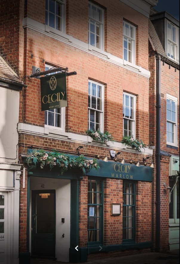 The exterior of Cecily Medi Spa features a brick façade with decorative greenery. A green sign with gold lettering displays 'Cecily Marlow.' Windows on the upper floors are framed with appealing details, indicating a welcoming and stylish entrance on the bustling street.