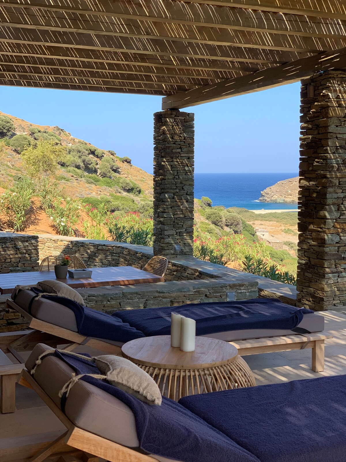 A shaded terrace is presented with comfortable loungers facing the expansive sea view. Natural stone columns and a rustic wooden table enhance the serene ambiance, while lush greenery and hills are visible in the background, creating a tranquil outdoor setting.