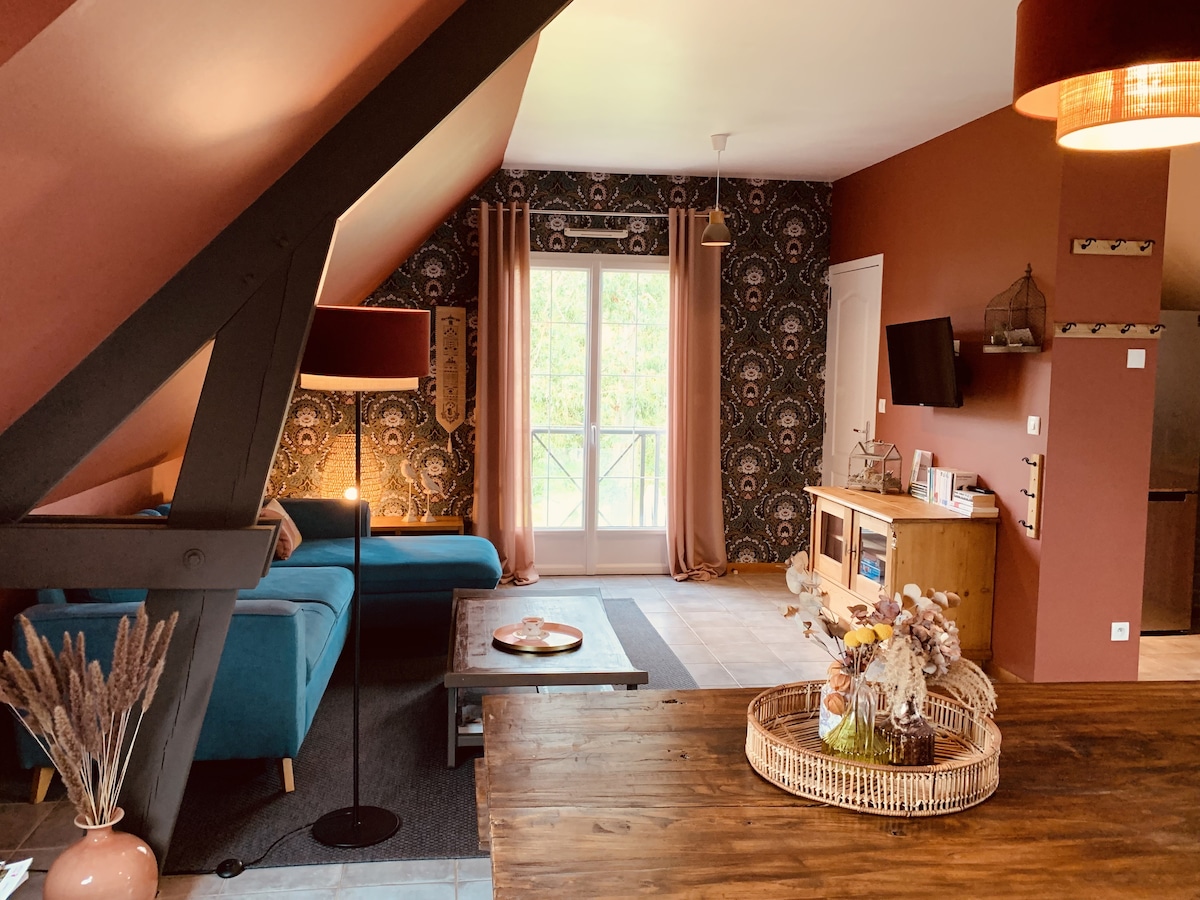 A spacious living area is presented with a blue sofa and wooden furnishings. Light streams through large windows adorned with beige curtains, illuminating the patterned wall. A decorative centerpiece with dried flowers is placed on the table, enhancing the room's cozy charm.