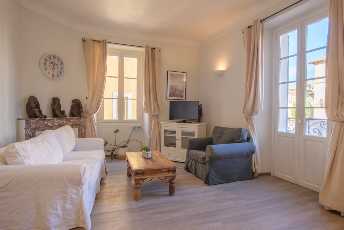 A well-lit living area includes a large window with French doors. A white sofa and a gray armchair are situated around a wooden coffee table, enhancing the room's comfort. Decorative sculptures and simple wall art provide a touch of character.