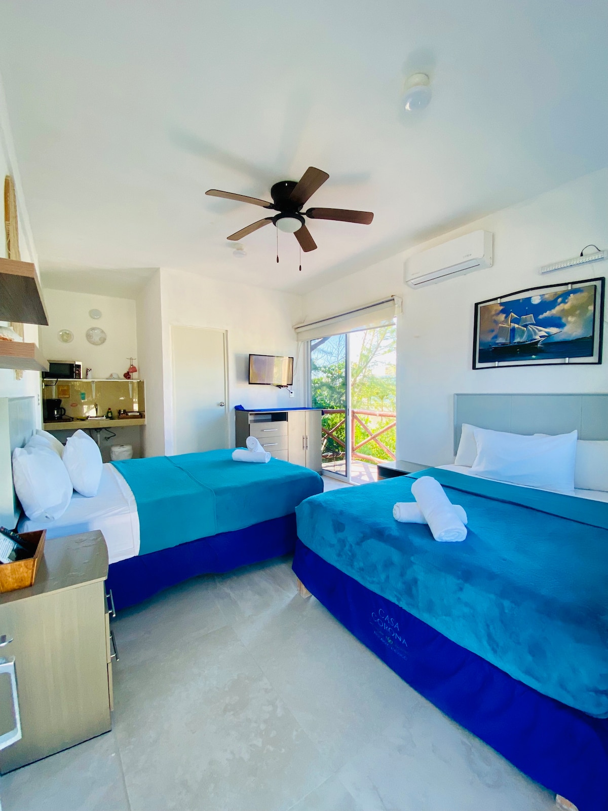 The room features two Queen Size beds, both adorned with soft blue blankets and neatly arranged towels. A compact kitchenette is visible in the background along with a television. Natural light flows in from the balcony door, showcasing a bright and airy atmosphere.
