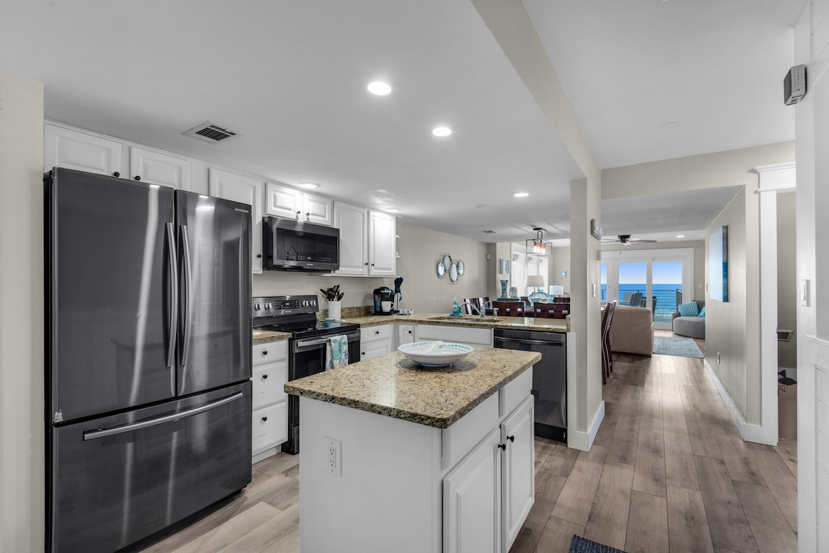 The kitchen features modern appliances, including a stainless steel refrigerator and a microwave. A large island with granite countertops serves as a functional space for cooking and dining. Ample cabinetry provides storage, while a connected dining area is visible in the background, offering ocean views.
