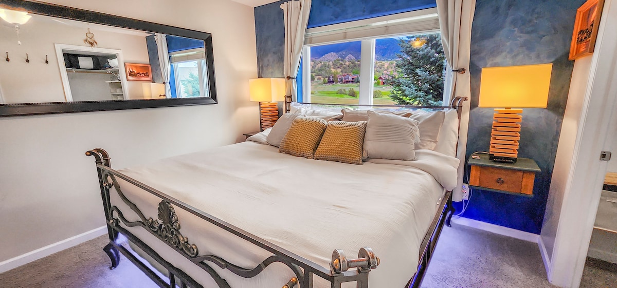 A spacious sleeping area features a large, inviting bed adorned with plush pillows. Soft, ambient light glows from bedside lamps, while a mirror reflects the serene colors of the room. A window offers views of the surrounding mountains and greenery, creating a calming atmosphere.
