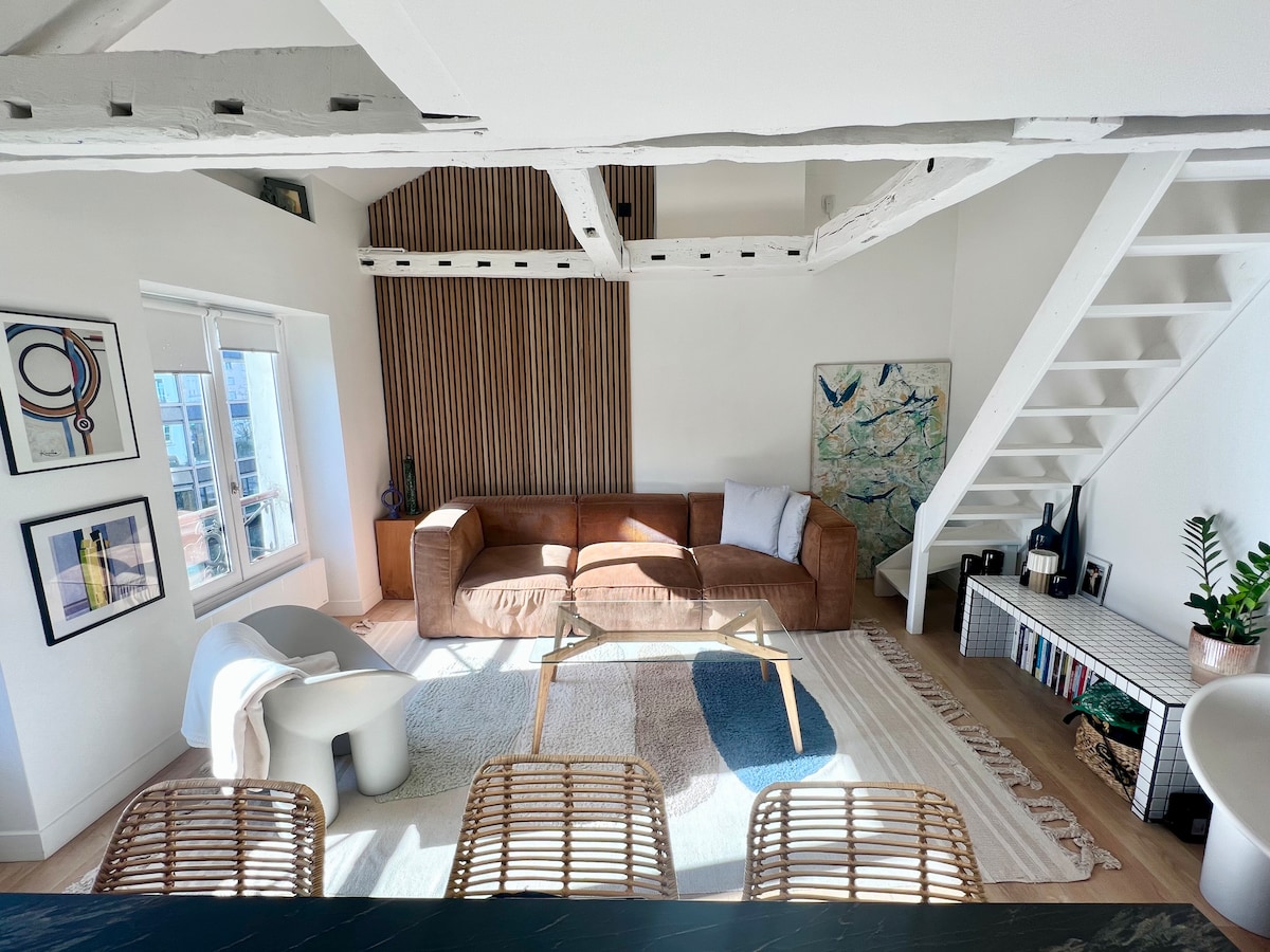A spacious living area is illuminated by natural light from a large window. Modern art pieces adorn the walls, while a comfortable brown sofa invites relaxation. A sleek coffee table is positioned on a soft area rug, with a staircase leading to the lofted sleeping area in the background.