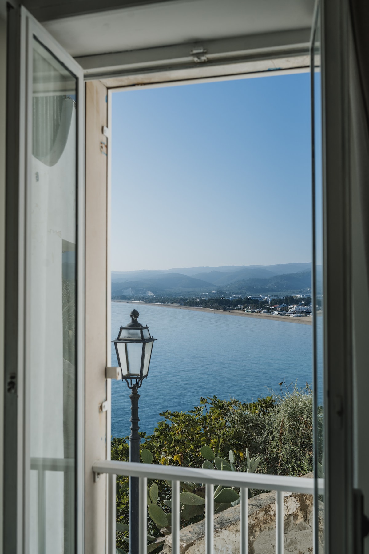 An open window reveals a stunning sea view, showcasing calm waters reflecting the blue sky. A vintage lantern stands along the shoreline, framed by lush greenery and distant hills, creating a serene and inviting setting.