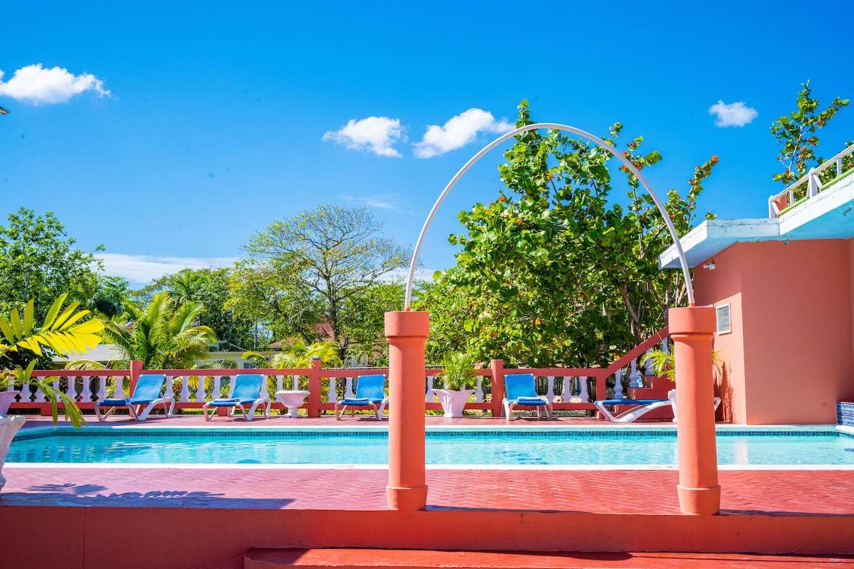 A clear blue sky frames a vibrant pool area surrounded by lush greenery. The pool features a unique archway, while lounge chairs are positioned along the edge. The surrounding architecture is painted in soft pastel colors, lending to a relaxed outdoor atmosphere.