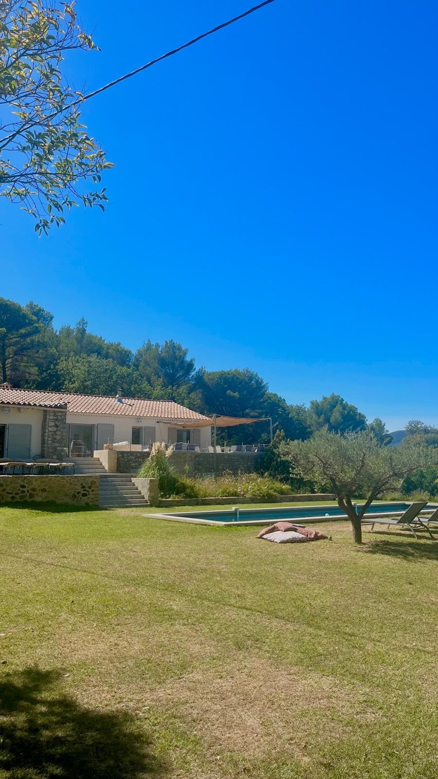 The exterior of the house features a light-colored facade and a covered terrace, flanked by vibrant greenery. A well-maintained lawn leads to a long pool, with loungers positioned nearby. The clear blue sky complements the tranquil outdoor setting.