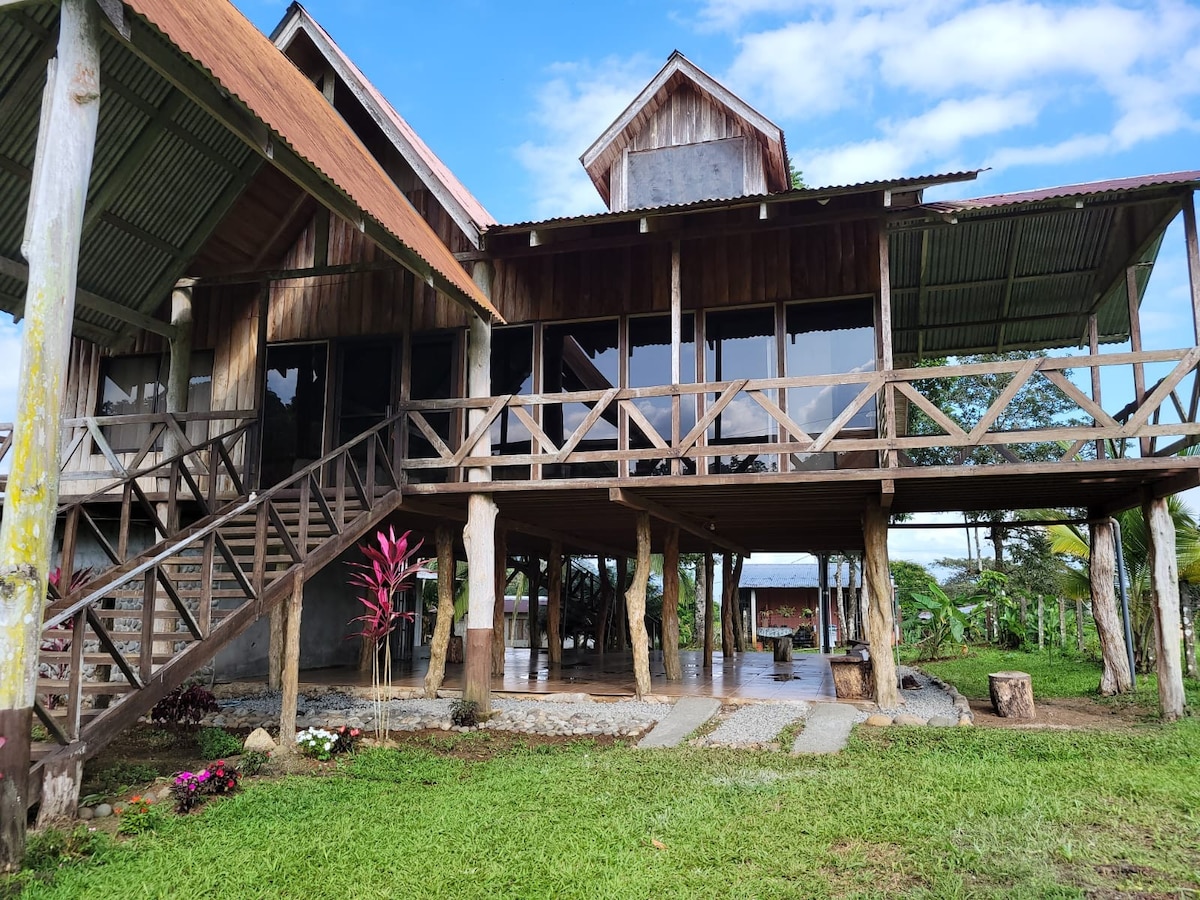 A spacious wooden house is elevated on stilts, featuring a large porch with a railing that overlooks the surrounding nature. Glass doors and windows provide views of the outdoors, while a stone pathway leads from the entrance to the lush green lawn.