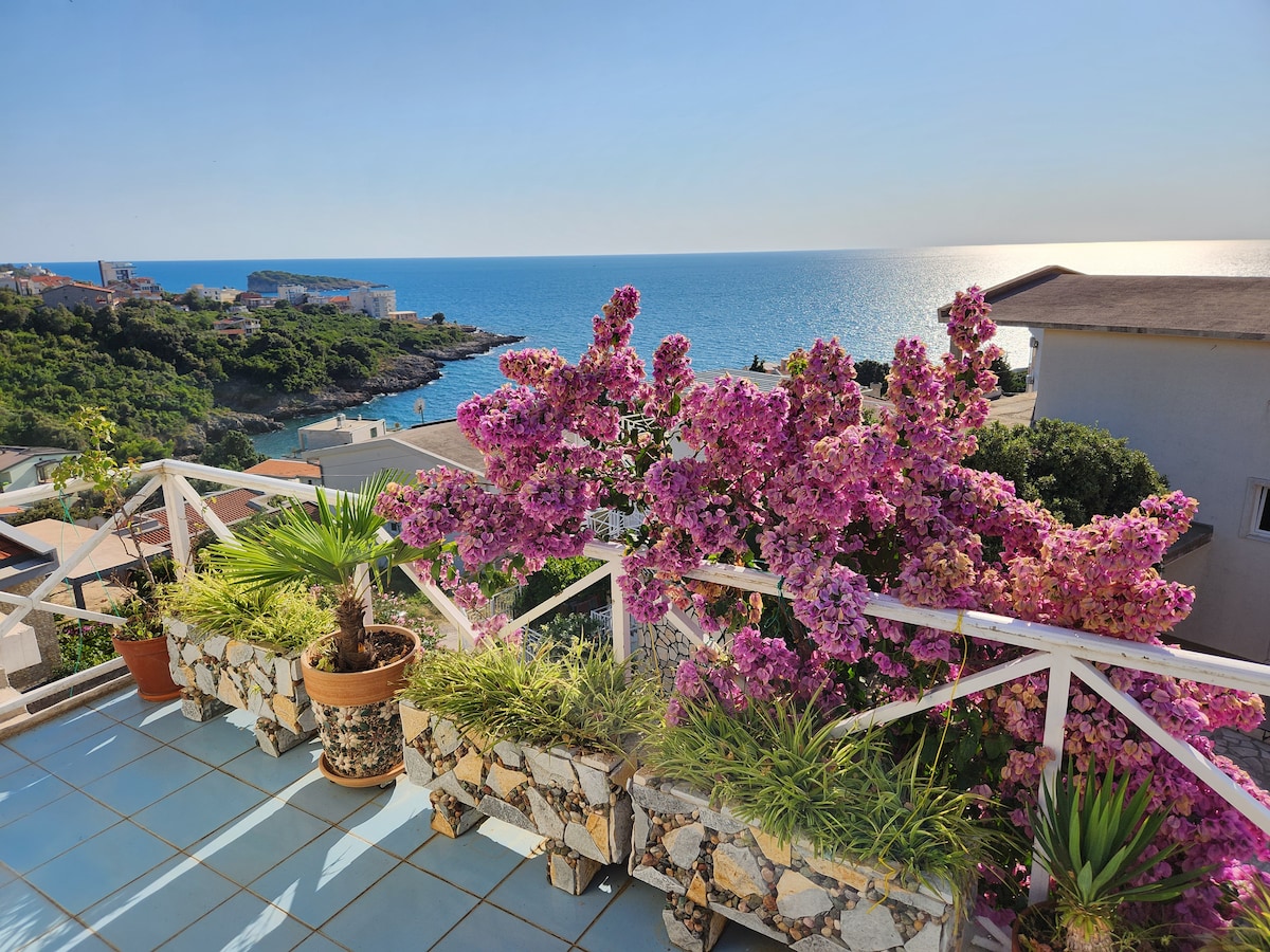 A balcony offers a panoramic view of the sea, bordered by vibrant flowering plants in pots. Lush greenery is visible alongside the colorful blooms, creating a serene environment for relaxation amidst the coastal scenery.