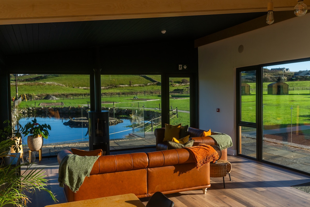 A spacious living area is highlighted by large glass windows offering views of lush, green fields. A brown leather sofa is complemented by decorative throws and a plant. Natural light fills the space, creating a bright, inviting atmosphere.