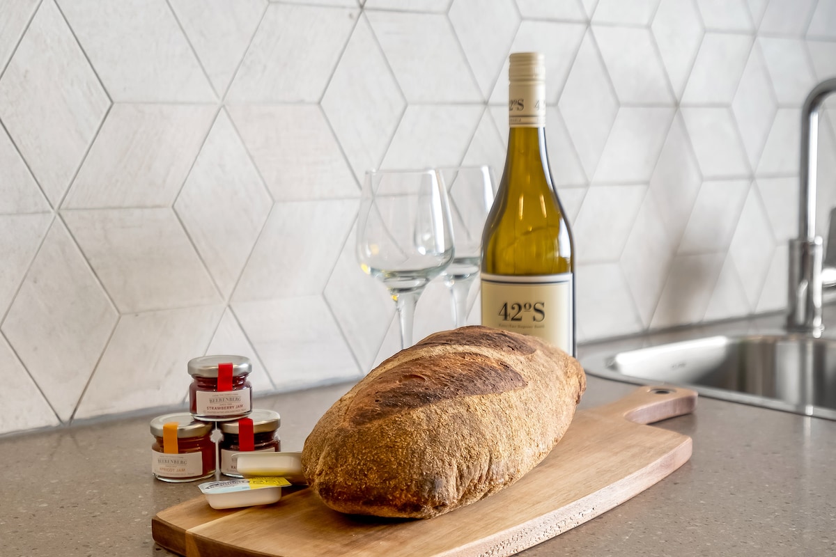 A wooden cutting board displays a freshly baked loaf of bread alongside jars of preserves and a butter dish. A bottle of white wine and two wine glasses are presented in the background, illuminated by natural light reflecting off the hexagonal tile wall.