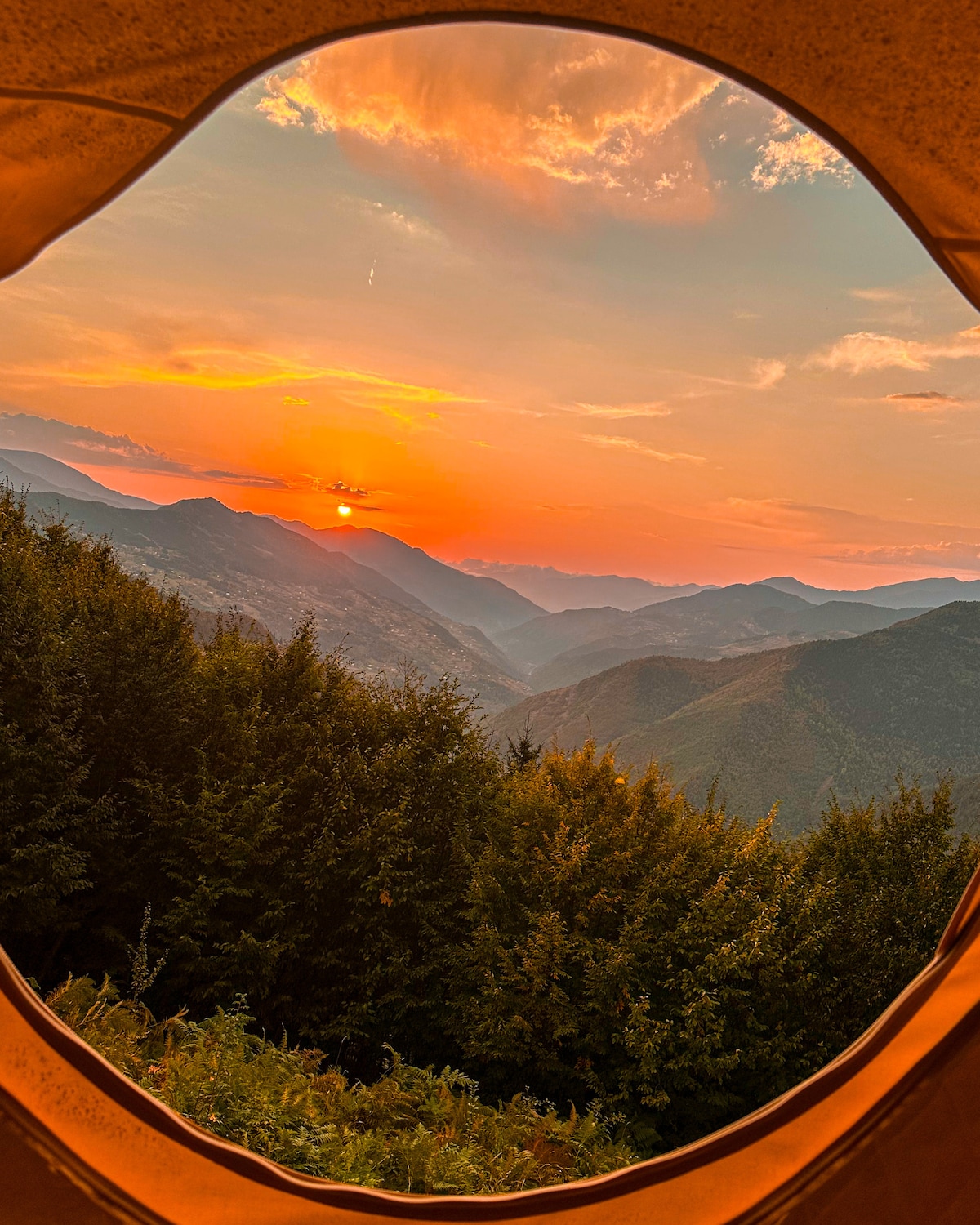 A view from inside the tent reveals a vibrant sunset over the mountains, framed by the round opening. Lush green trees and hills create a serene backdrop, enhancing the natural beauty of the landscape.