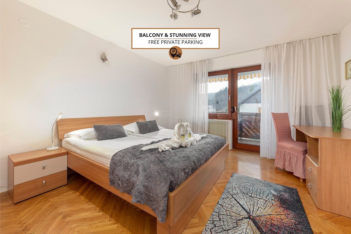 A spacious bedroom is featured, equipped with a large bed and a decorative throw. Natural light enters through glass doors leading to a private balcony, revealing mountain views. A desk and a chair complement the room, while light curtains softly frame the windows.