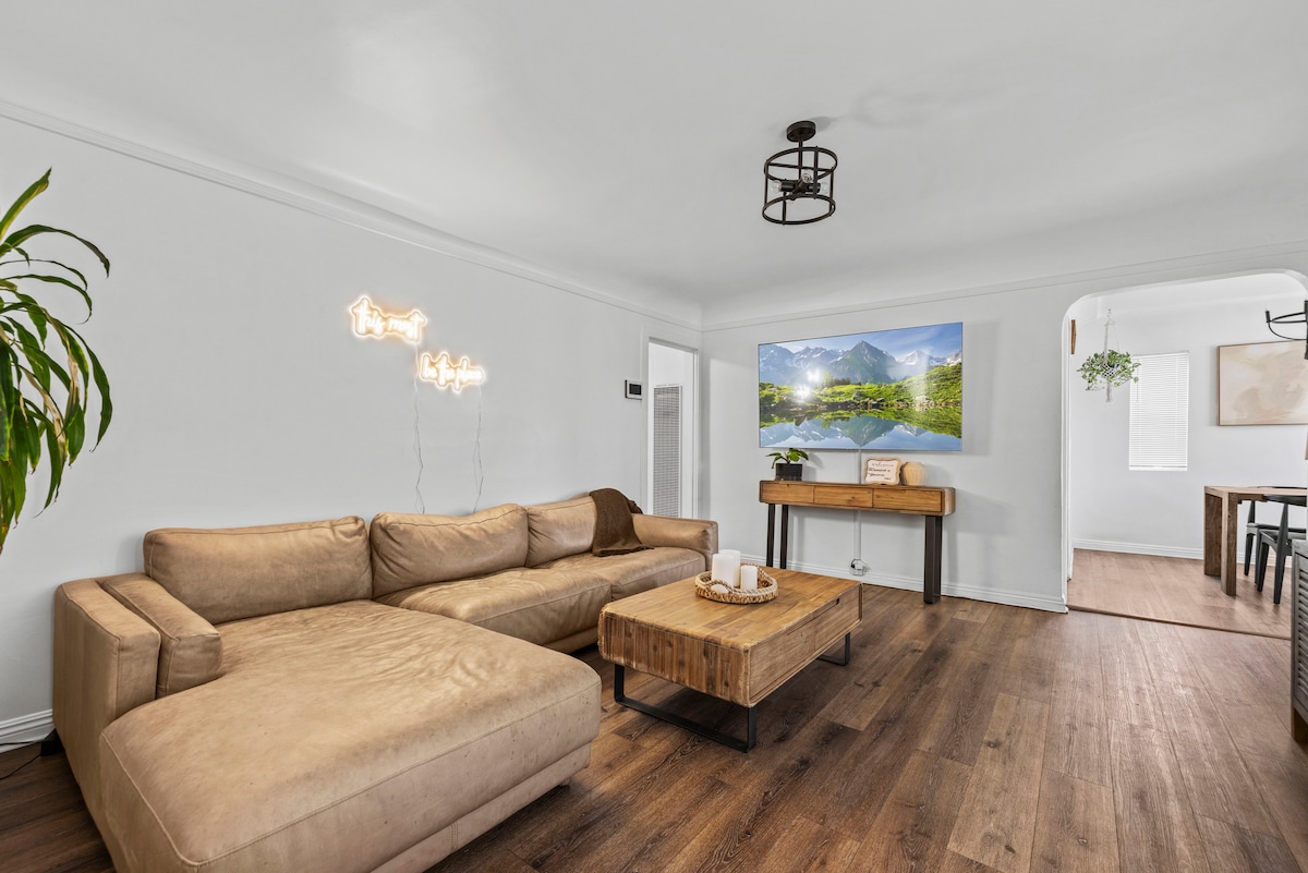 A spacious living room is highlighted by a large sectional couch and a wooden coffee table. A large wall-mounted TV displays a serene mountain scene. Natural light is welcomed through a nearby window, enhancing the bright, neutral color palette.