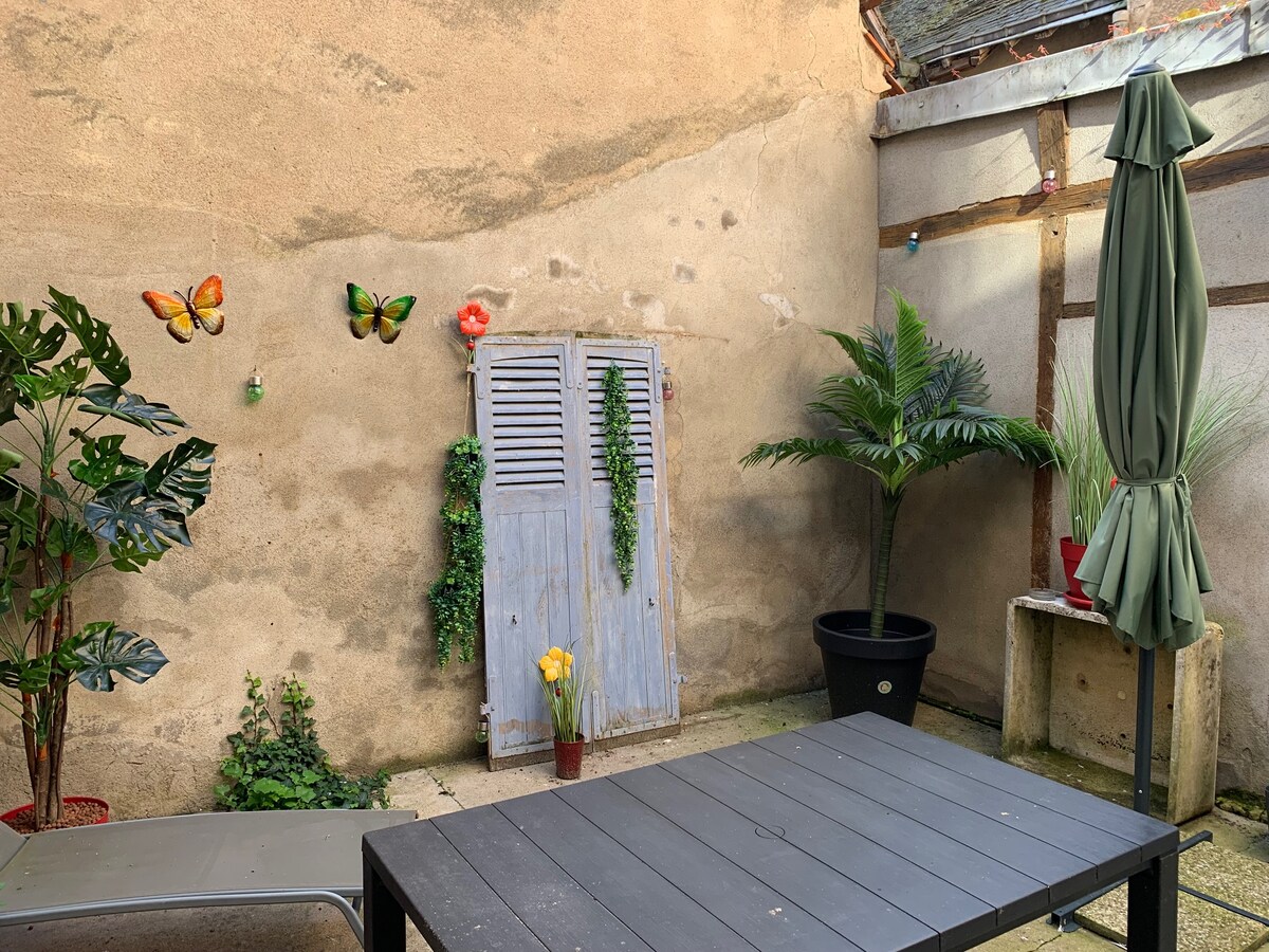 A quaint patio area is featured, with a wooden table and lounge chairs positioned on stone tiles. Decorative butterflies adorn the wall, alongside a blue shutter and potted greenery. A large umbrella offers shade, while flowers add a touch of color to the space.