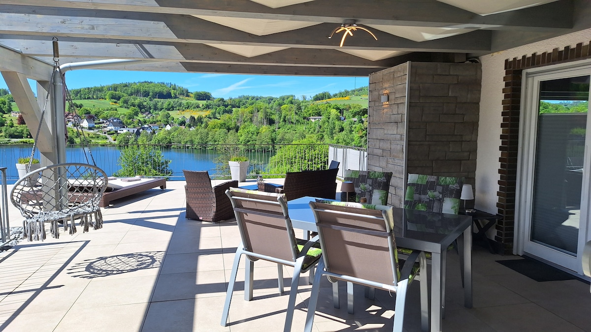 A spacious terrace features comfortable seating arrangements, including a dining table and lounge chairs, set against a backdrop of lush greenery and a serene lake view. Natural light floods the area, highlighting the well-appointed outdoor furniture under a shaded roof.