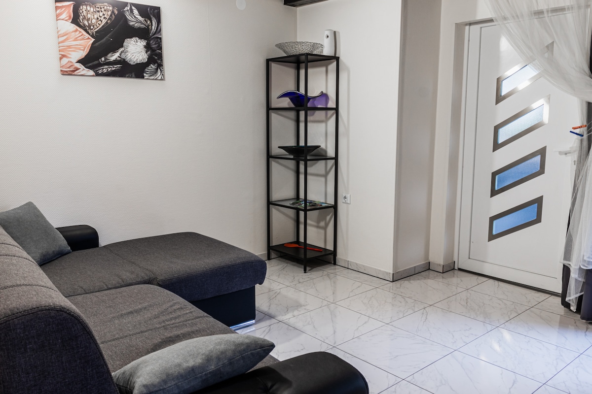 A modern living area features a dark gray sectional sofa with cushions, positioned next to a minimalist black shelving unit displaying decorative items. A light-colored door with geometric glass panels provides natural light, while the glossy tile floor reflects the room's layout.