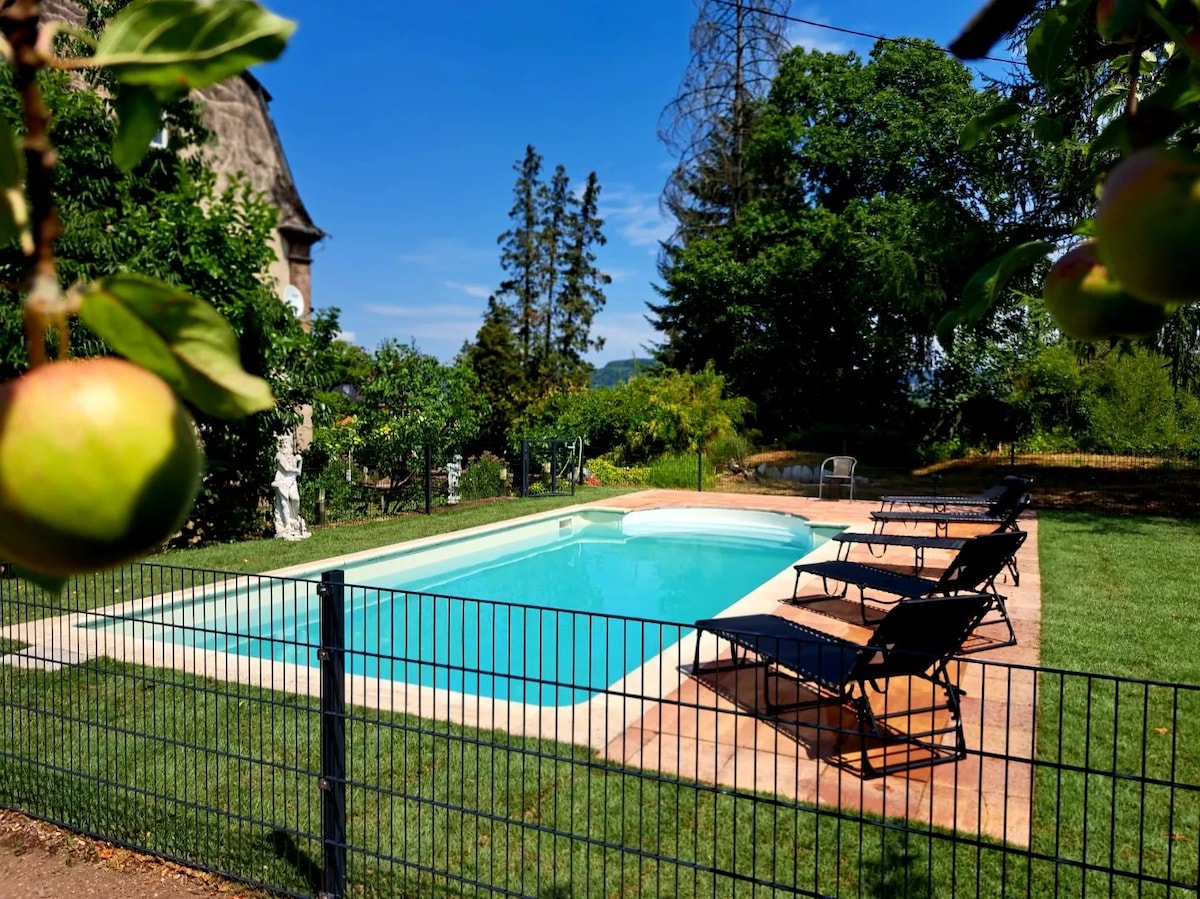 A fenced swimming pool area features crystal clear water surrounded by a stone patio. Sun loungers are arranged neatly along the edge. Lush greenery and trees provide a tranquil setting, with a glimpse of the castle structure visible in the background.
