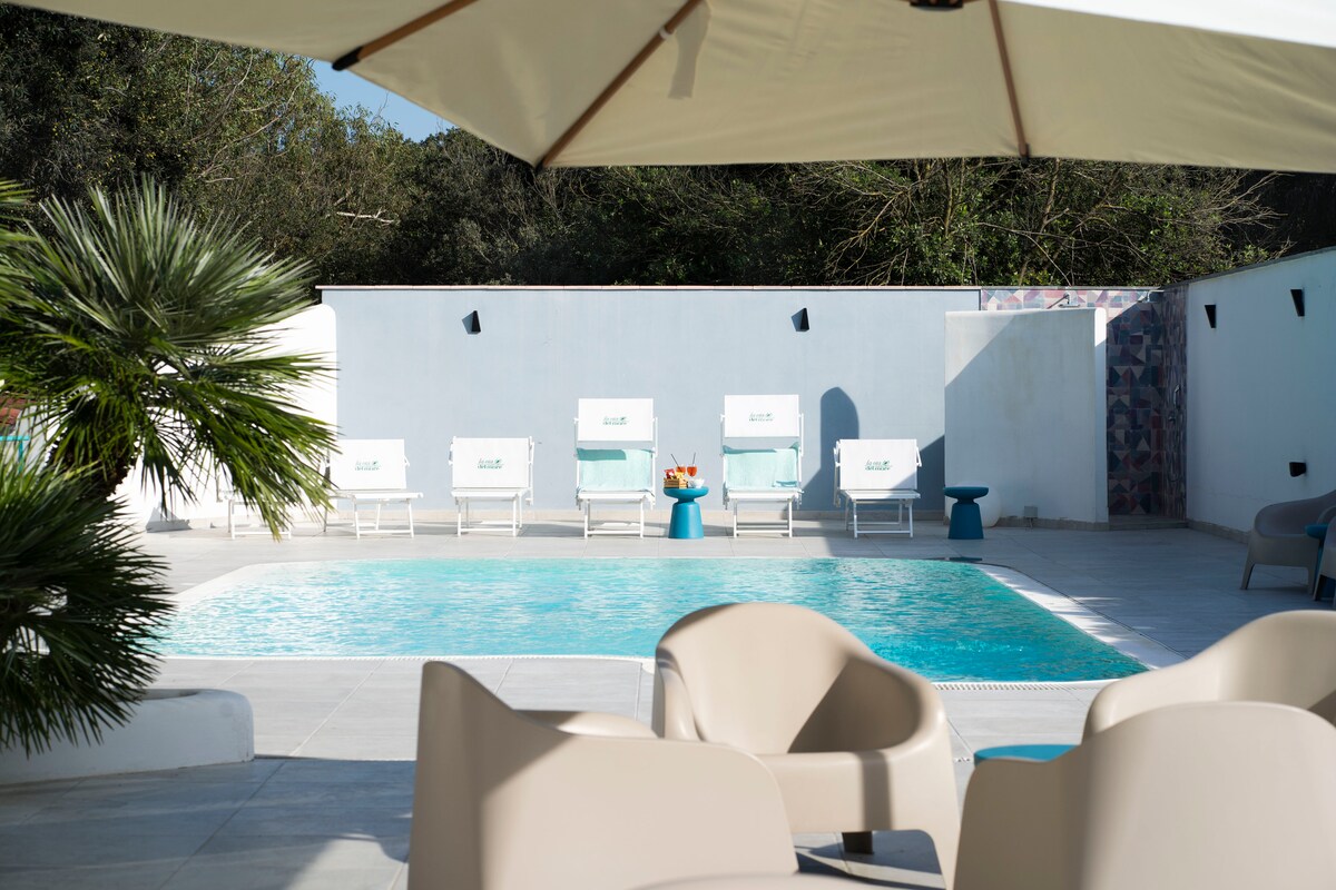 An inviting outdoor space features a clear swimming pool surrounded by lounge chairs. A series of white sun loungers are arranged near the water, complemented by a tropical plant. The area is shaded by a large umbrella, enhancing the tranquil setting.