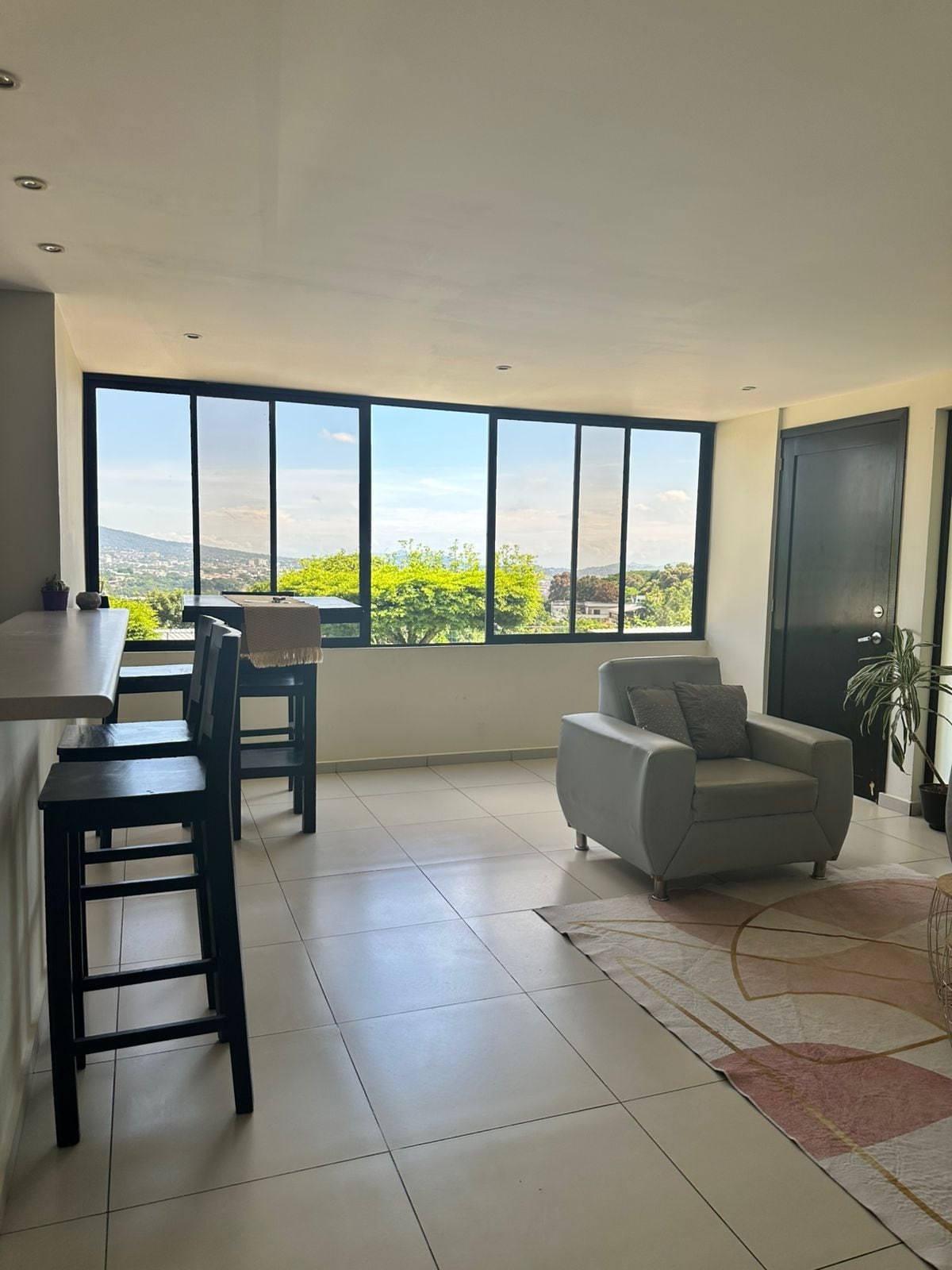 A bright and airy living space features large windows providing a view of the surrounding greenery. A small armchair sits on a patterned area rug. Tall chairs are positioned near a bar counter, creating an inviting atmosphere for relaxation or socializing.