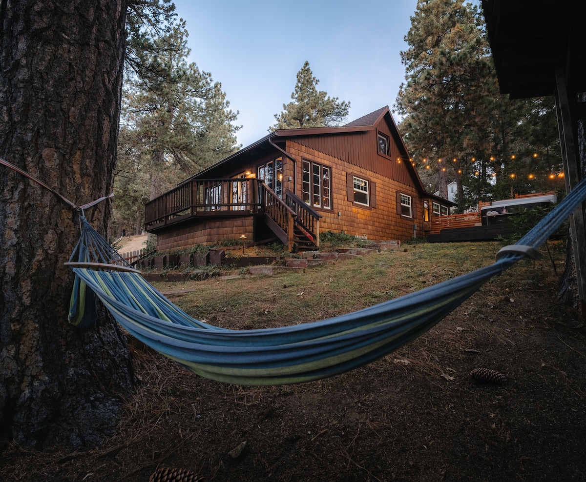 A vintage cabin surrounded by tall trees is depicted, featuring a spacious deck with string lights. A colorful hammock is shown hanging between two trees in the foreground, inviting relaxation. The warm glow from the cabin's windows suggests a cozy atmosphere within.