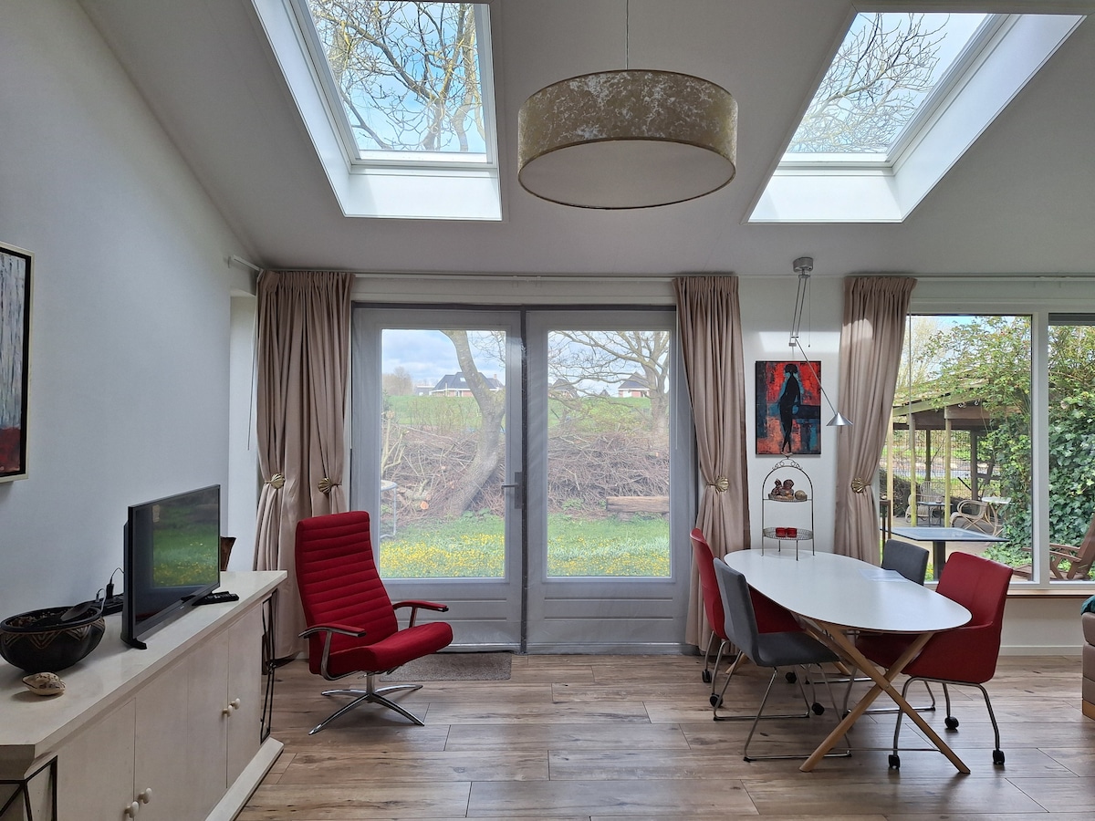 The light-filled living space features large skylights and expansive glass doors that offer a view of the lush garden. A round dining table is surrounded by red chairs, while a red armchair adds a pop of color. Natural wood flooring complements the warm ambiance.