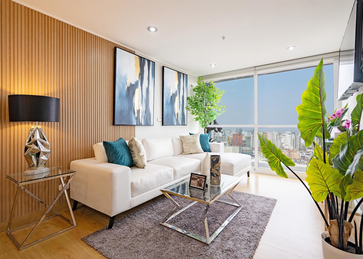 A modern living area features a white sectional sofa positioned against a wooden accent wall. Two large paintings adorn the wall, complemented by a green potted plant. A glass coffee table sits on a soft area rug, with natural light flooding through the expansive window.