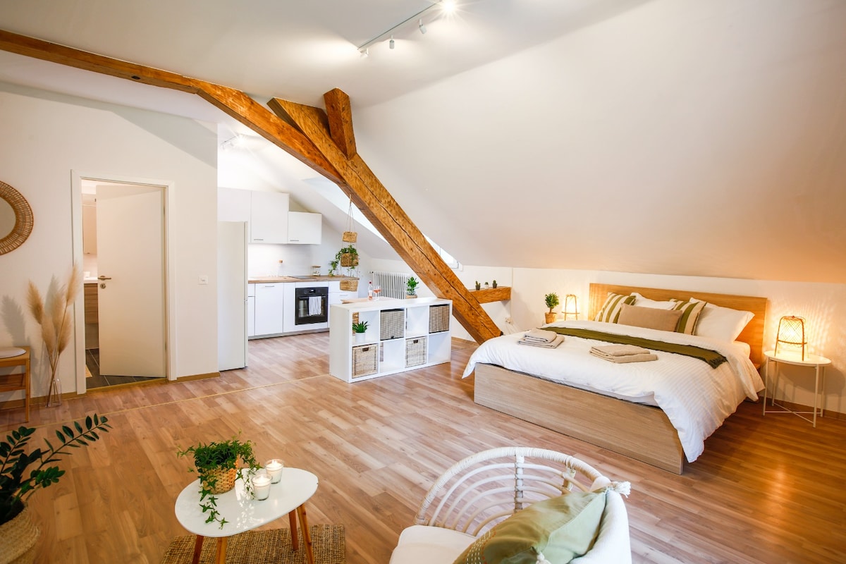 The spacious studio features exposed wooden beams and a natural color palette. A comfortable bed is positioned next to soft lighting and minimalist decor. A small dining area is visible, with a fully equipped kitchen in the background, enhancing the open-plan layout.