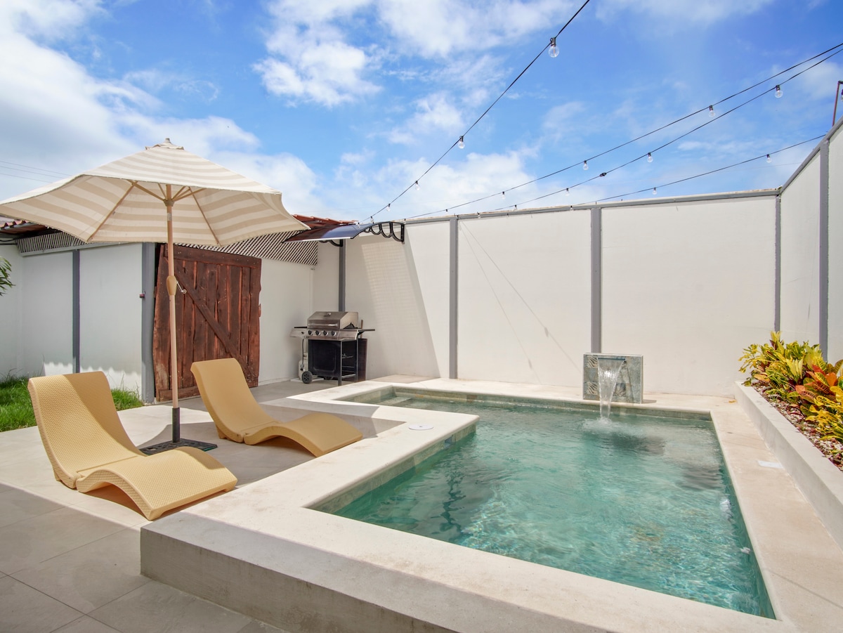 A private pool area features a rectangular pool with a serene water fountain, surrounded by beige tiles. Two lounge chairs are positioned nearby under a decorative umbrella. String lights are installed overhead, enhancing the outdoor relaxation space.