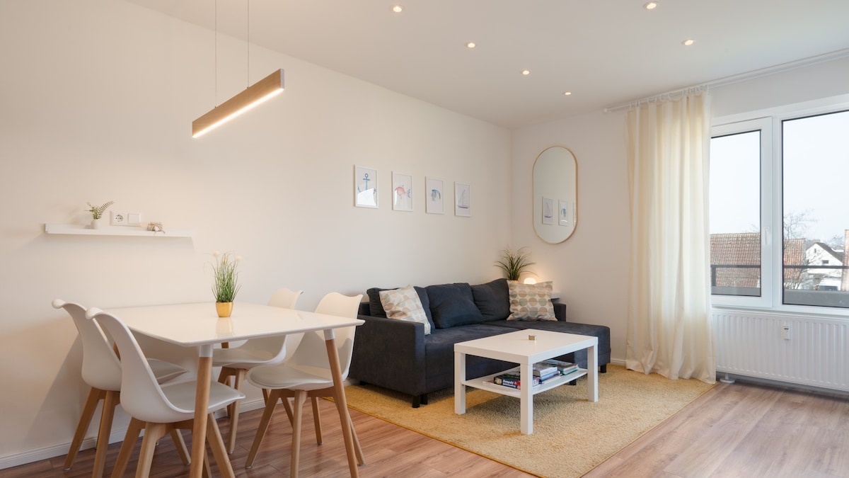 A modern living area is featured, showcasing a dark sofa against a light backdrop. A white dining table with chairs occupies one side, while a coffee table sits in front of the sofa. Natural light enters through large windows, complemented by soft curtains.