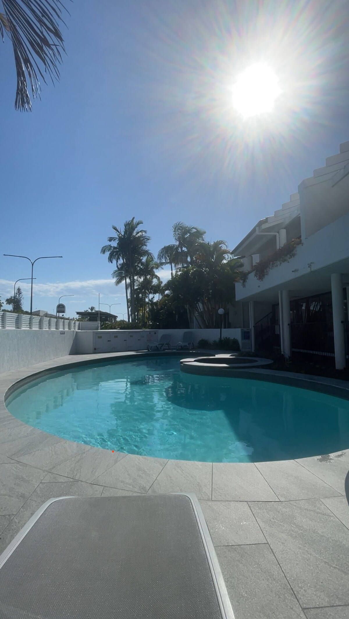 A circular pool is presented, surrounded by a neatly paved area and tropical palm trees. Sunlight reflects off the water's surface, enhancing the inviting appeal. The upper levels of the complex are visible in the background, along with outdoor furniture positioned around the pool for relaxation.