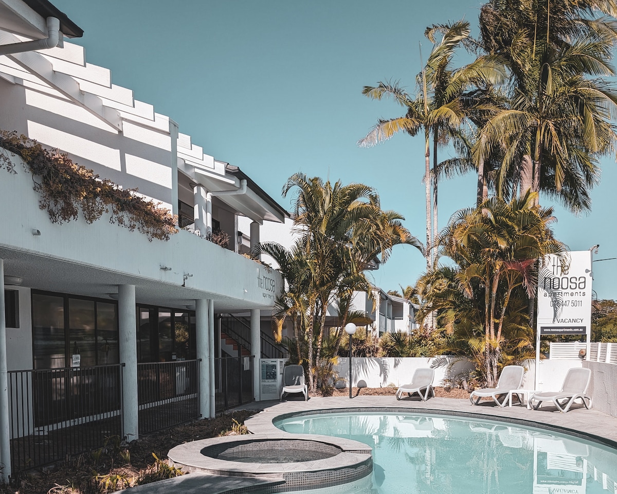 A refreshing pool area is bordered by palm trees and tropical landscaping. Sun loungers are arranged nearby, providing a relaxing outdoor space. The building features a modern façade with large windows that invite natural light, enhancing the overall vibrant atmosphere.
