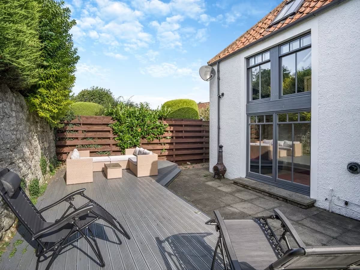 An outdoor decking area is shown, furnished with light-colored seating and reclining loungers. The space is surrounded by greenery and features a stone pathway leading to the house, which is characterized by large windows allowing ample natural light.