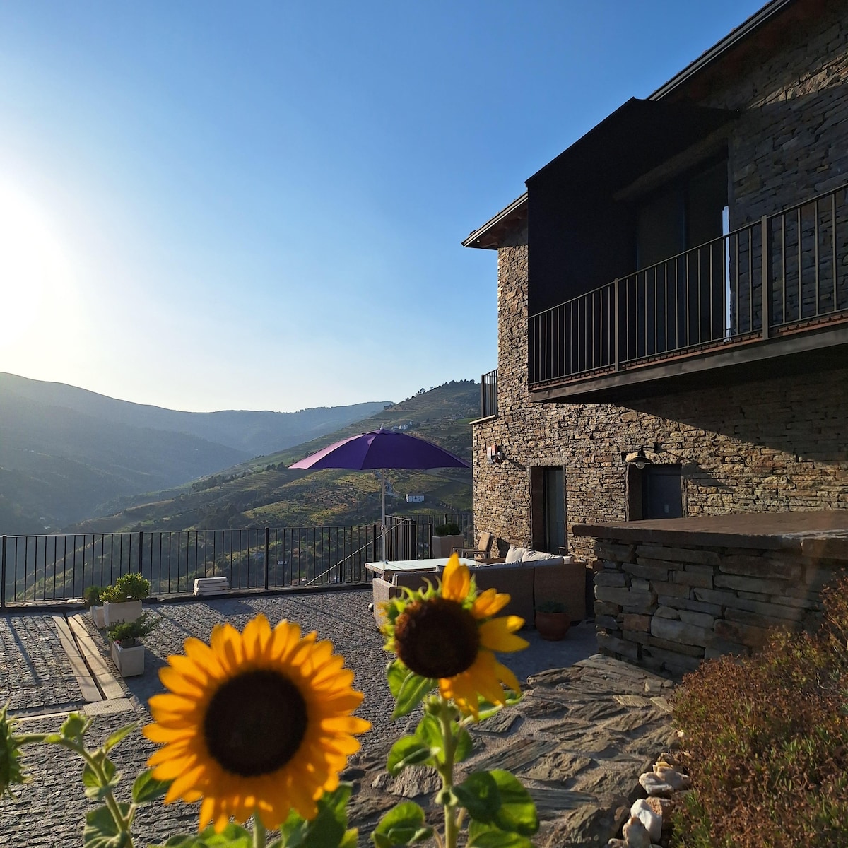 A charming stone exterior is showcased, complemented by vibrant sunflowers in the foreground. A purple umbrella provides shade over a cozy seating area, with breathtaking mountain views and a clear sky enhancing the serene atmosphere.