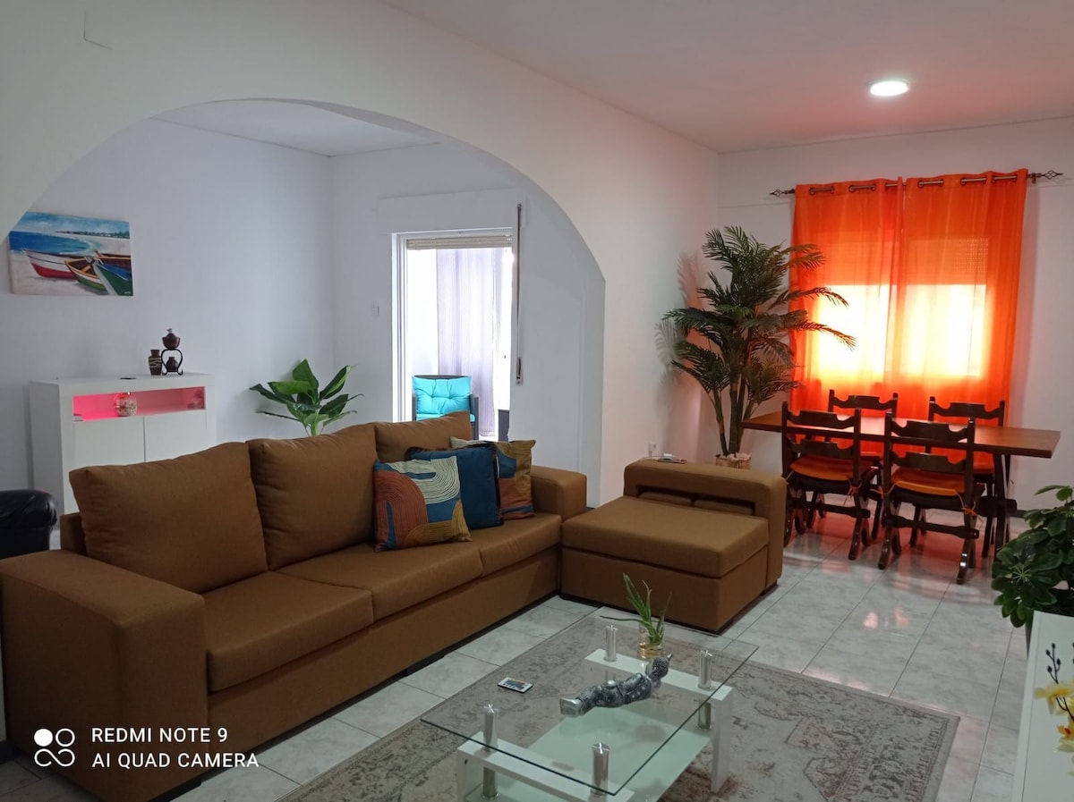 The living area features a spacious brown sofa with an adjoining chaise lounge, accompanied by a glass coffee table. Bright orange curtains frame a window, while a dining table with chairs sits in the background, and green potted plants add a touch of greenery.