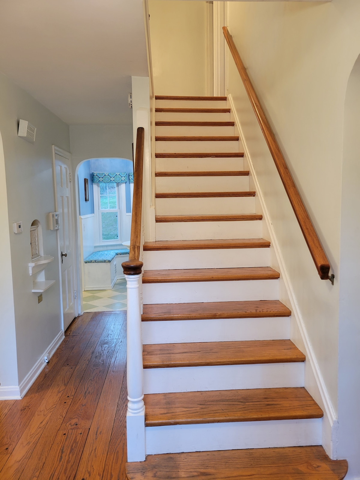 A staircase with wooden steps leads to the upper floor. The handrail is finished in natural wood. A hallway is visible on the left, with a nook featuring a cushioned seat, while light pours in through a nearby window.