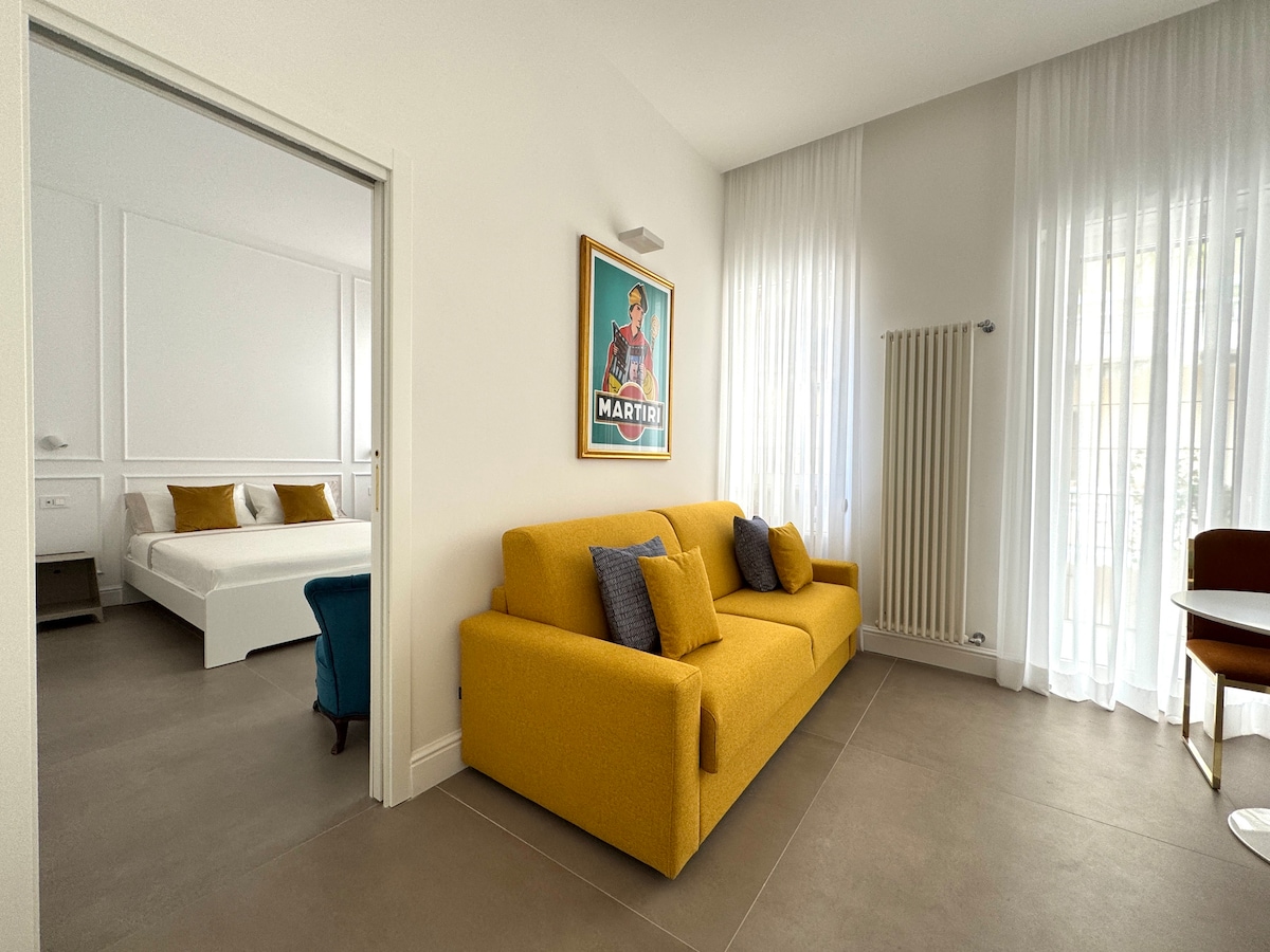 A bright living area features a yellow sofa that complements the neutral decor. Behind the sofa, a spacious bedroom can be seen, with a large bed and light-colored linens. Natural light filters through sheer curtains, enhancing the room's airy atmosphere.