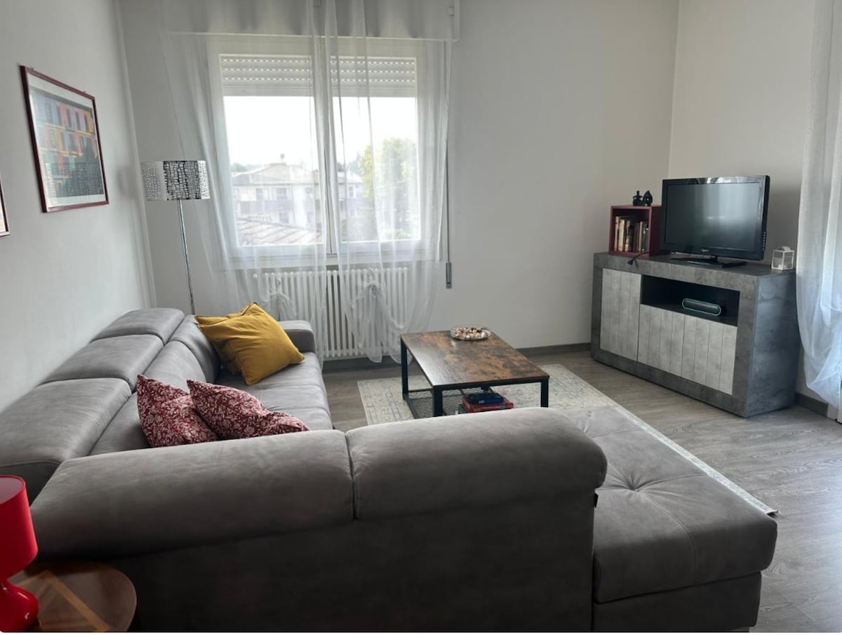 A spacious living area features a comfortable grey sectional sofa adorned with decorative cushions and a bright yellow throw. A wooden coffee table sits in front, complemented by a modern TV console and natural light entering through large windows dressed with sheer curtains.