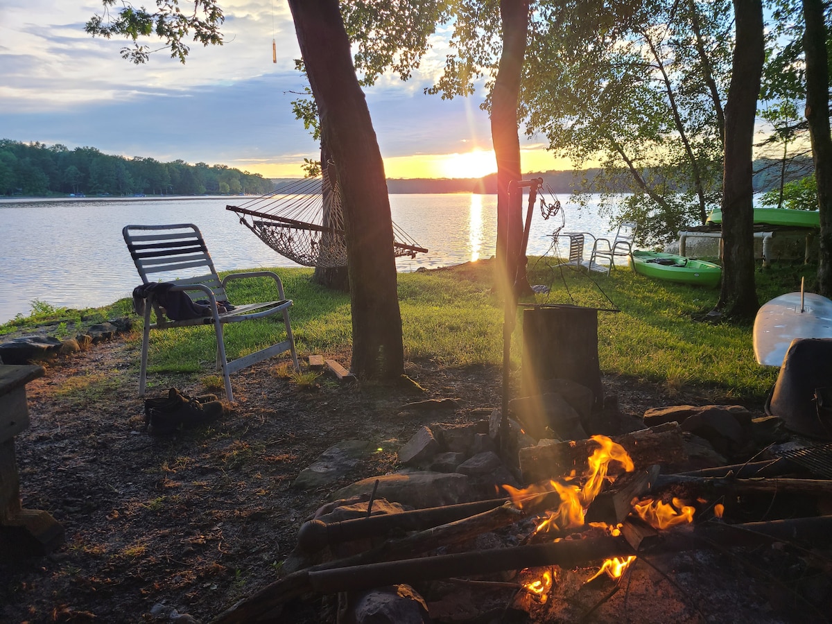 The lakeside view features a warm campfire in the foreground, surrounded by stones. A hammock hangs between trees nearby, with kayaks resting on the shore. Soft lighting from the sunset reflects on the water, creating a tranquil atmosphere.