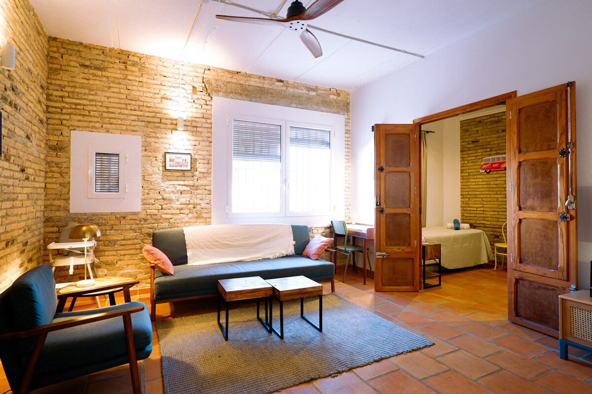 A spacious living area features exposed brick walls and rustic wooden doors. Comfortable seating options include a sofa and armchairs, arranged around two wooden coffee tables on a textured rug. Natural light enters through the window, enhancing the room's inviting ambiance.