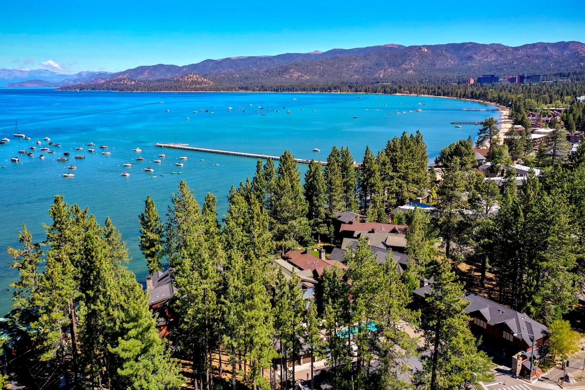 A scenic view captures the tranquil waters of Lake Tahoe, dotted with boats. Lush green trees frame the shoreline, while the distant mountains add depth to the landscape under a clear blue sky.