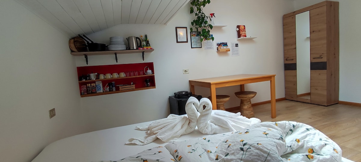The room features a spacious bed adorned with a floral duvet cover and towel swans. A wooden table with two stools is positioned beside a red shelf displaying kitchenware. A wardrobe stands against the wall, with a light-colored wooden floor completing the cozy atmosphere.