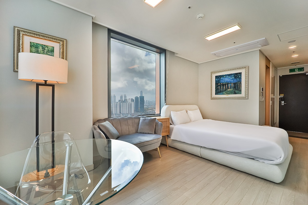A spacious room features a queen size bed with soft bedding and a scenic city view visible through the large window. A modern glass dining table and a cozy seating area with a stylish couch complement the layout, creating a comfortable atmosphere.