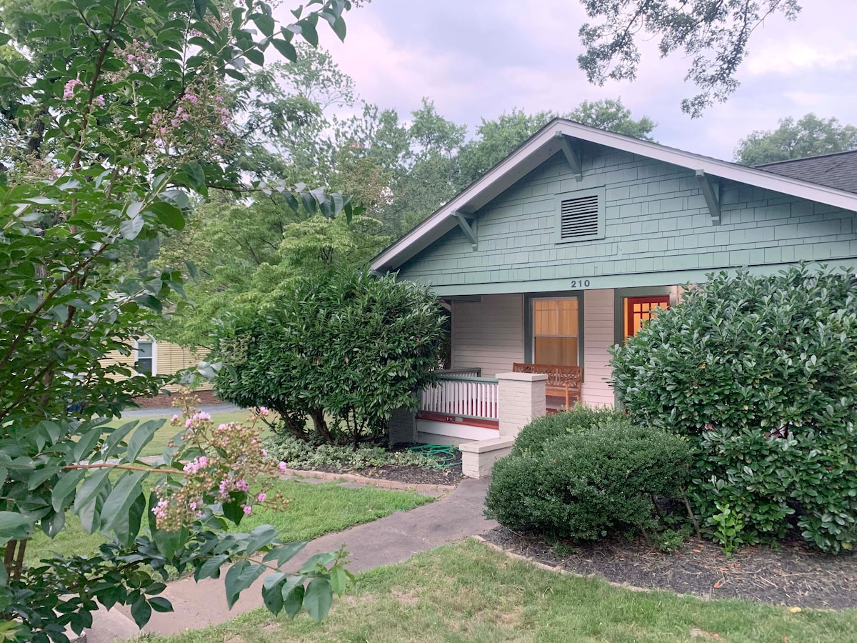 The exterior of a craftsman-style cottage is showcased among lush greenery. A welcoming front porch is highlighted, featuring wooden rocking chairs. The pathway is lined with flowering plants, leading to the entrance. The home is set against a backdrop of trees, enhancing its serene appearance.