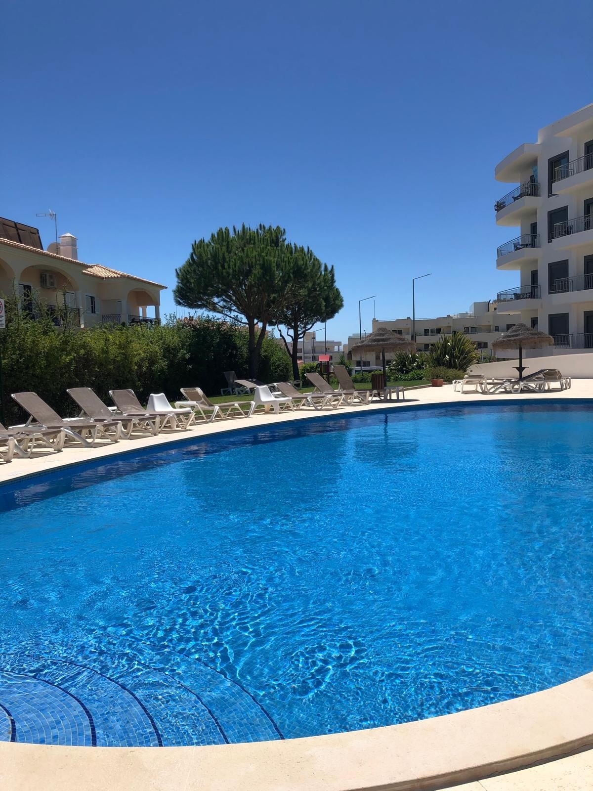 A clear blue swimming pool is framed by lounge chairs, inviting relaxation. Lush greenery is visible in the background, enhancing the outdoor space. The modern building stands nearby, providing a contemporary feel to the setting.