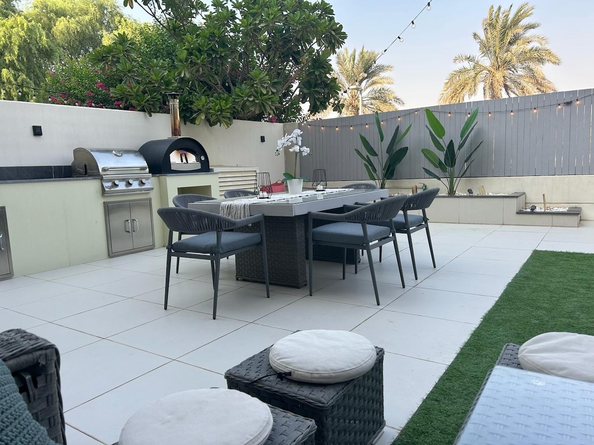 An outdoor dining area showcases a spacious table surrounded by several chairs, set against a backdrop of greenery. A built-in BBQ and pizza oven are present, with warm string lights adding gentle illumination. Paved flooring leads to a small patch of artificial grass.