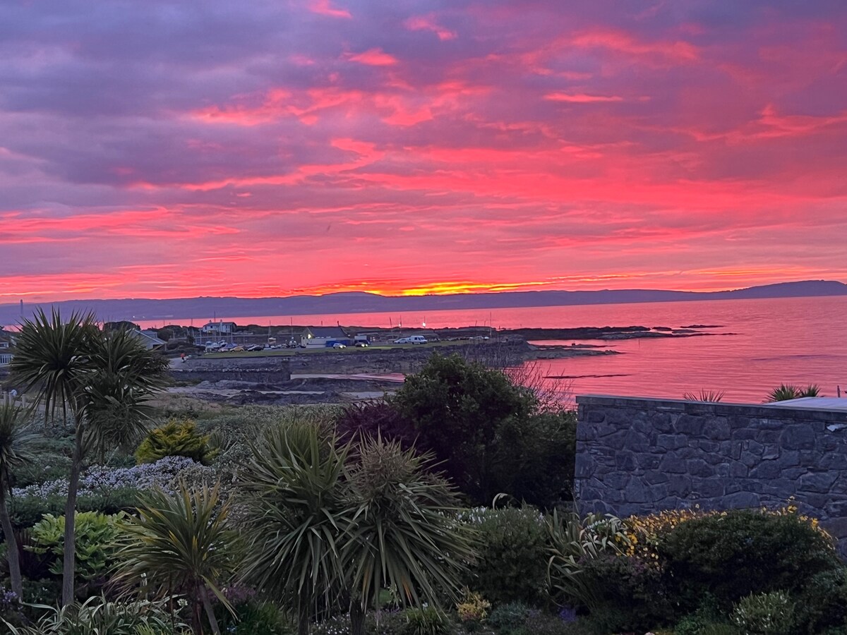 A breathtaking sunset casts vibrant hues across the sky, reflecting on the tranquil ocean waters. The coastline is visible in the foreground, flanked by lush greenery and colorful plants, creating a serene atmosphere that complements the natural beauty of the setting.